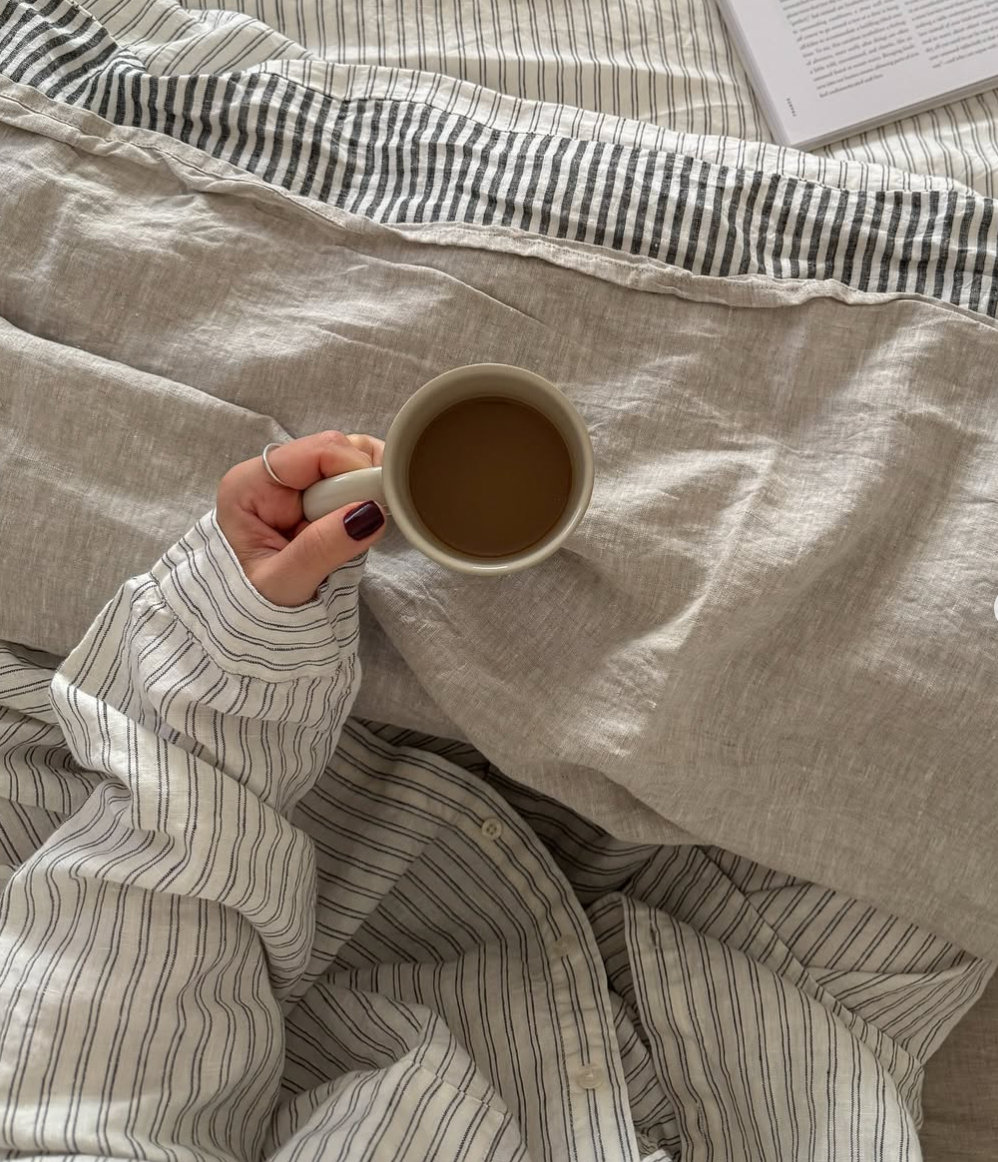 Simple Linen Co. 100% French flax linen sheet set in Nature featured with Ida in charcoal stripe and double pinstripe, paired with double pinstripe night to daywear long set, model drinking coffee in bed cozy bed, top down view.