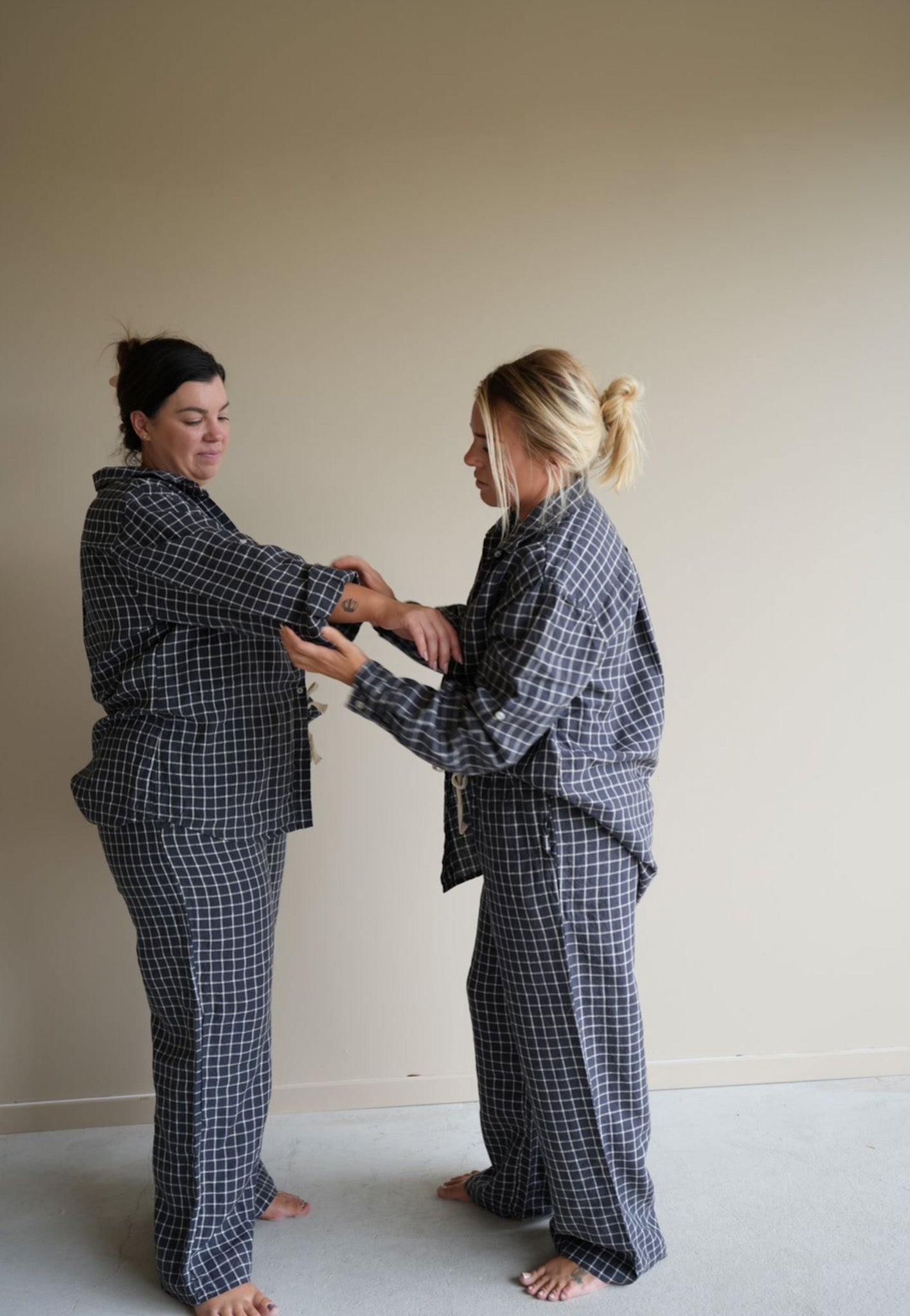 One model adjusting another’s sleeve, both wearing Simple Linen Co’s 100% French Flax Linen day to night wear long sleeve set in Navy Grid. 